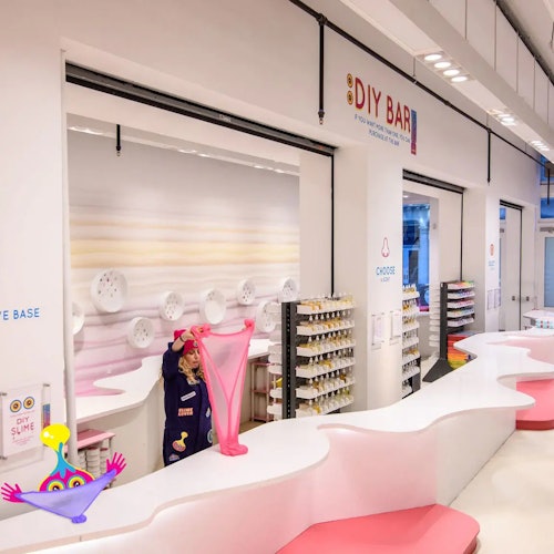 Un niño estira slime rosa en un mostrador de un bar de manualidades con estanterías llenas de materiales de creación, en un espacio moderno y bien iluminado.