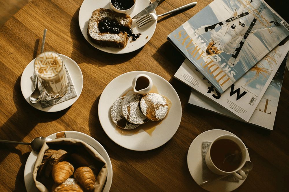 Pancakes, toast, and coffee on a wooden table beside a stack of ski magazines
