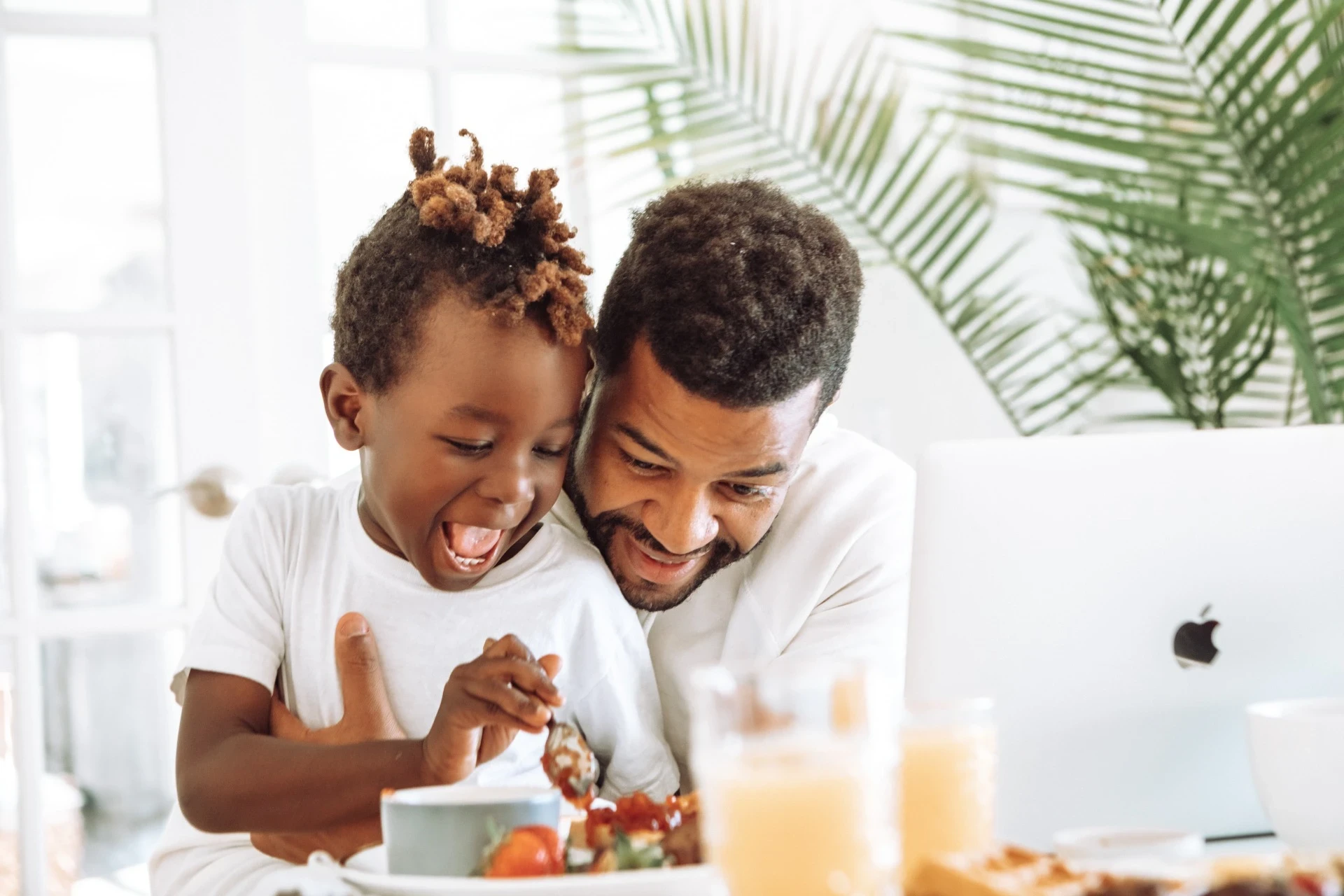 A imagem mostra um momento alegre entre um pai e seu filho, sentados juntos à mesa. Ambos estão sorrindo e aparentam estar aproveitando a refeição ou um lanche. O pai, de barba e com expressão carinhosa, envolve o filho em um abraço caloroso, enquanto o menino parece muito animado, segurando uma colher. Ao fundo, há um laptop com o logotipo da Apple, sugerindo que o pai pode estar trabalhando ou estudando enquanto passa um tempo com seu filho. A cena transmite afeto, felicidade e momentos de conexão em família, com uma ambientação clara e cheia de luz natural.