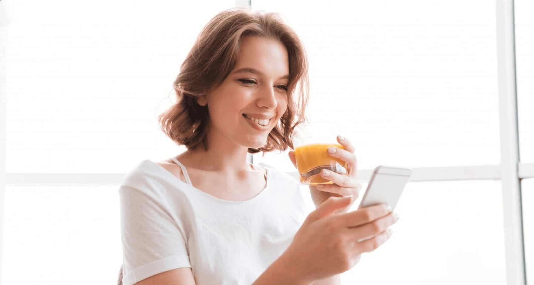 A young woman drinking juice and using her mobile phone, smiling and engaged with the device