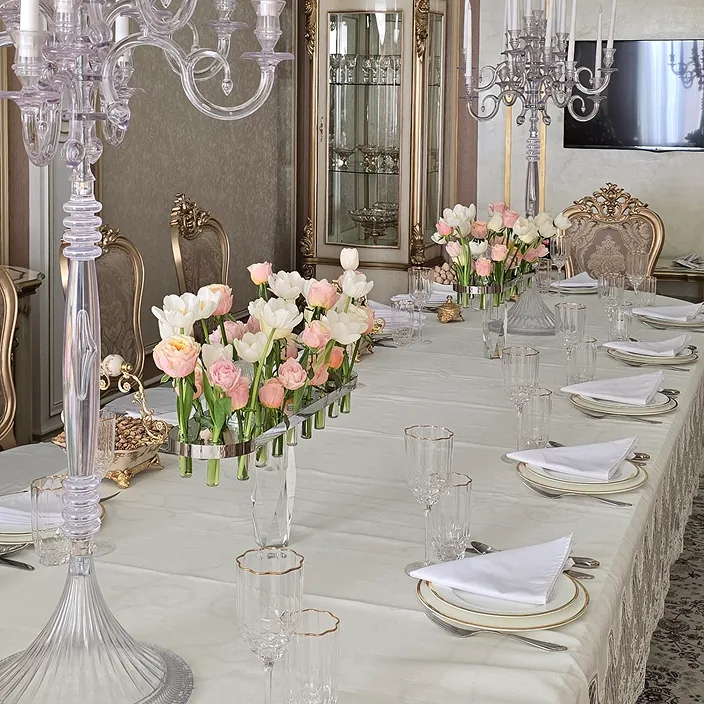 Formal dining table with white tablecloth, gold-rimmed plates, crystal glassware, and pink-white tulip centerpieces in glass vases, surrounded by ornate gold chairs in a classical, luxurious room.