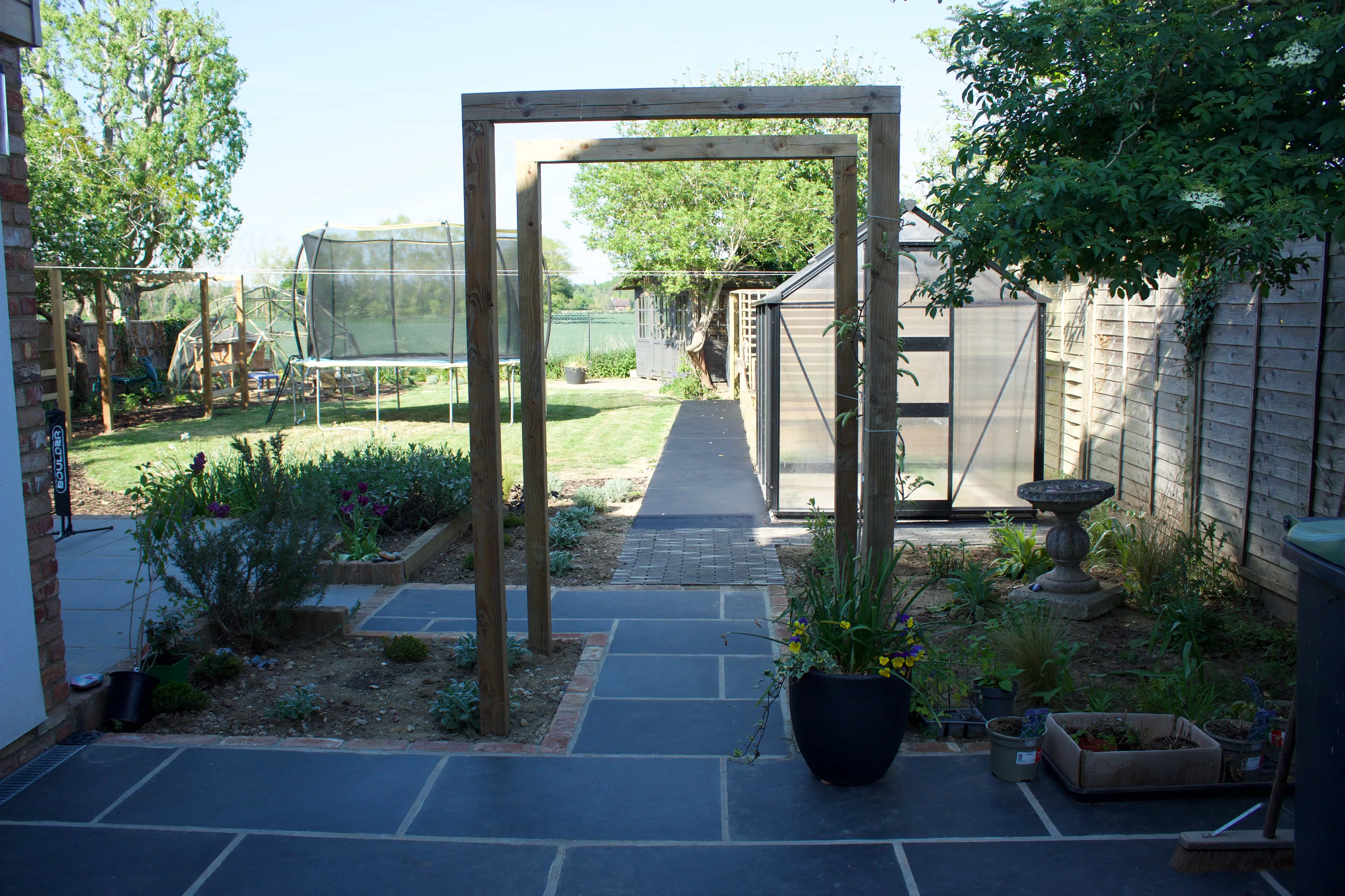 A pathway framed by a wooden archway, lined with plants and trees, leading to a grassy area.