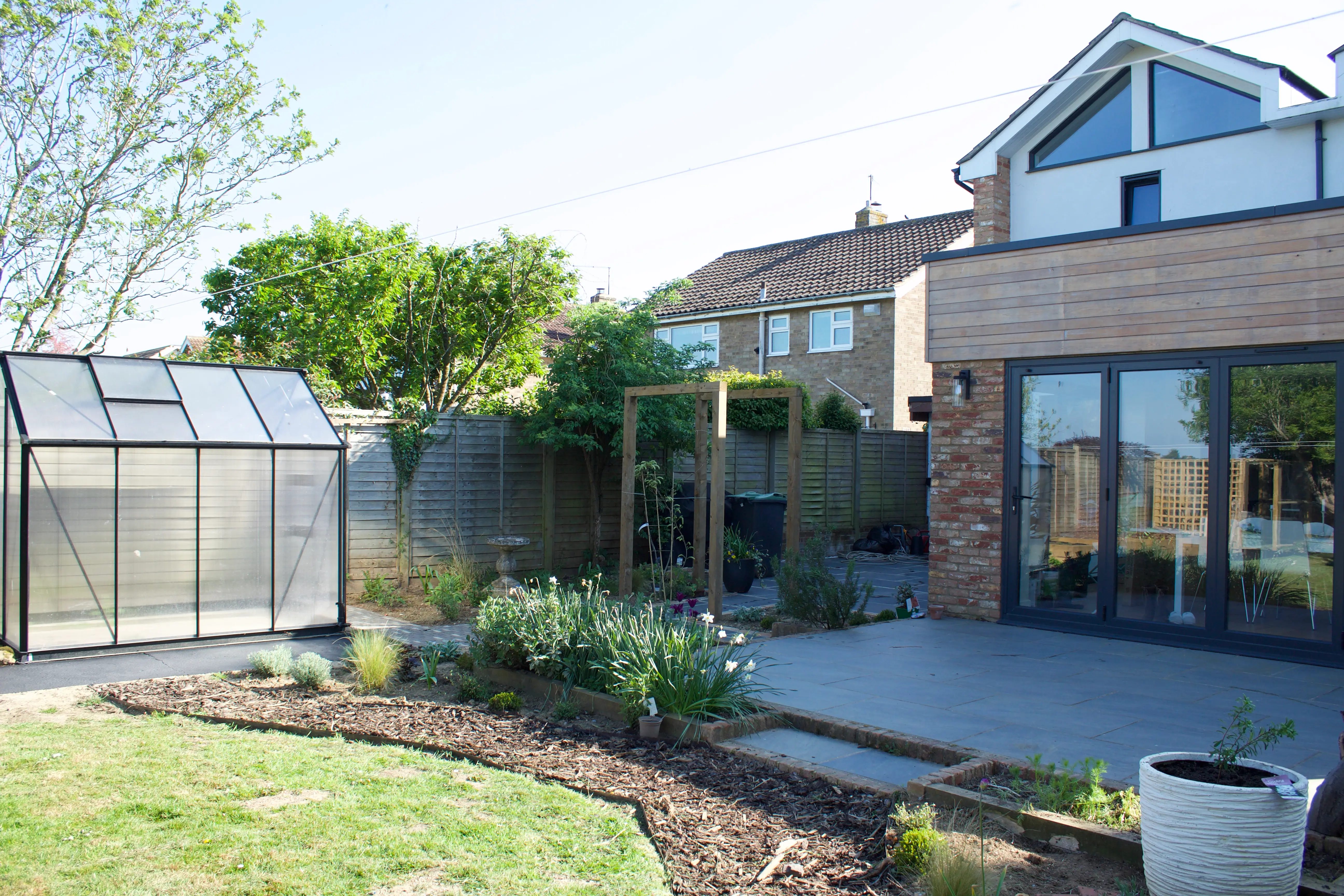 A sunny backyard with a house, garden beds, and a wooden fence, featuring a shed and lush greenery.