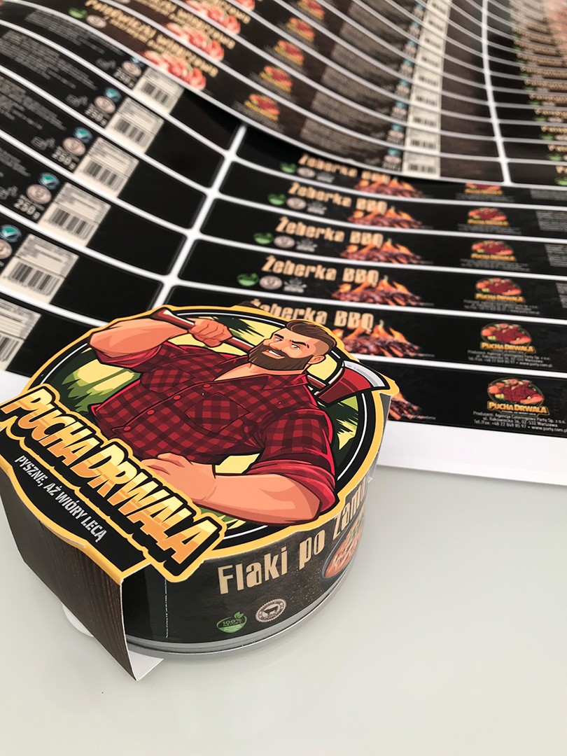 Rows of colorful packaging labels for "Pucha Drwala" are displayed on a flat surface, featuring vivid graphics of grilled meat and tin cans, highlighting their branding and nutritional information.