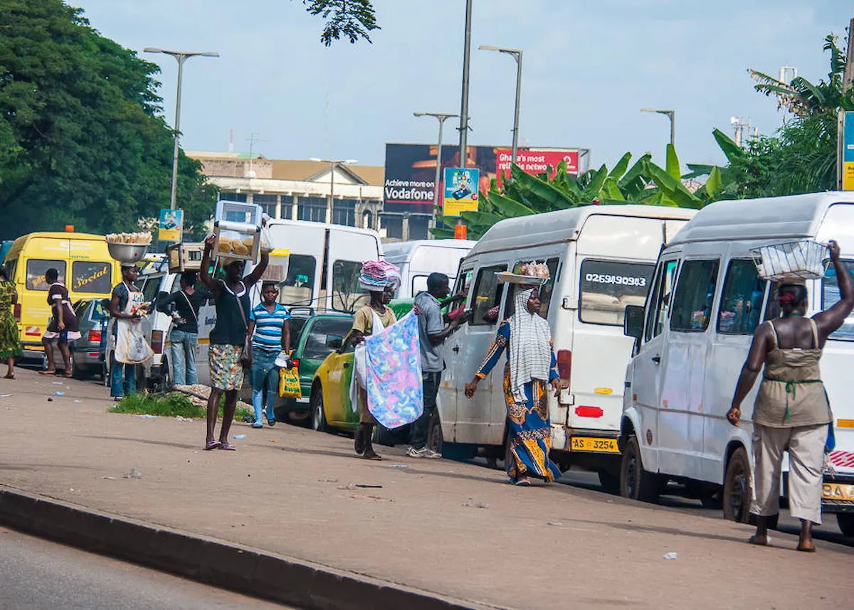 Busy Kumasi street with vendors and trotros at a transport hub.