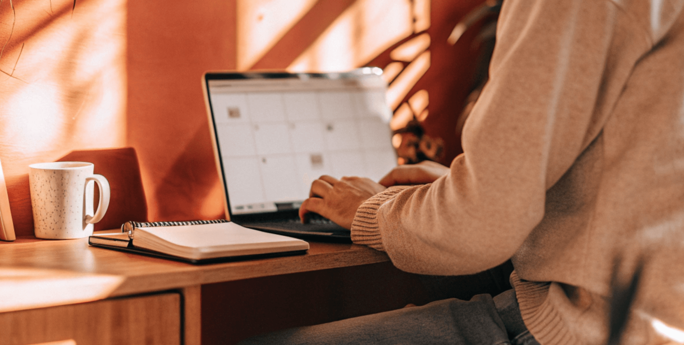 Person working on a laptop at a wooden desk with a coffee mug and notebook.