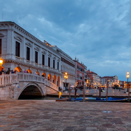 Veduta serale di un canale a Venezia con un ponte in pietra, lampioni accesi ed edifici storici. Le persone passeggiano lungo la passeggiata.