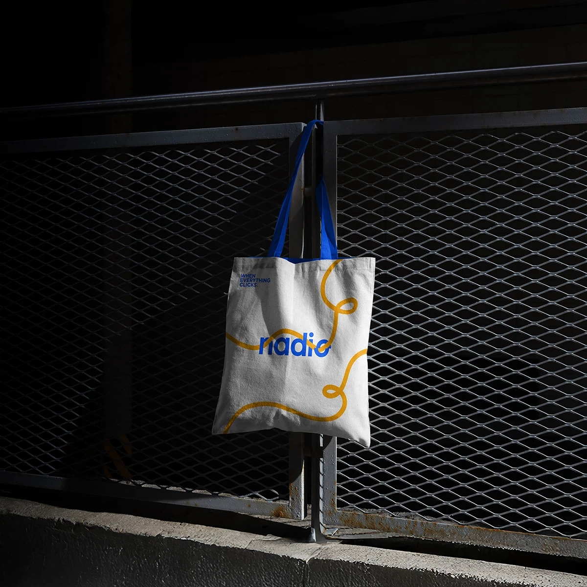 Tote bag with blue handles and yellow designs, featuring the word "radio" on a metal fence.