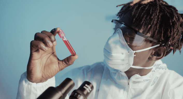 A laboratory researcher wearing a white protective suit, safety goggles, and a face mask holds up a red-filled test tube for close examination, with a microscope visible in the foreground.