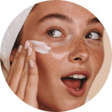 A person with a towel-wrapped hair applies moisturizer to their cheek, displaying healthy skin and a surprised expression; relaxed, skincare, routine.