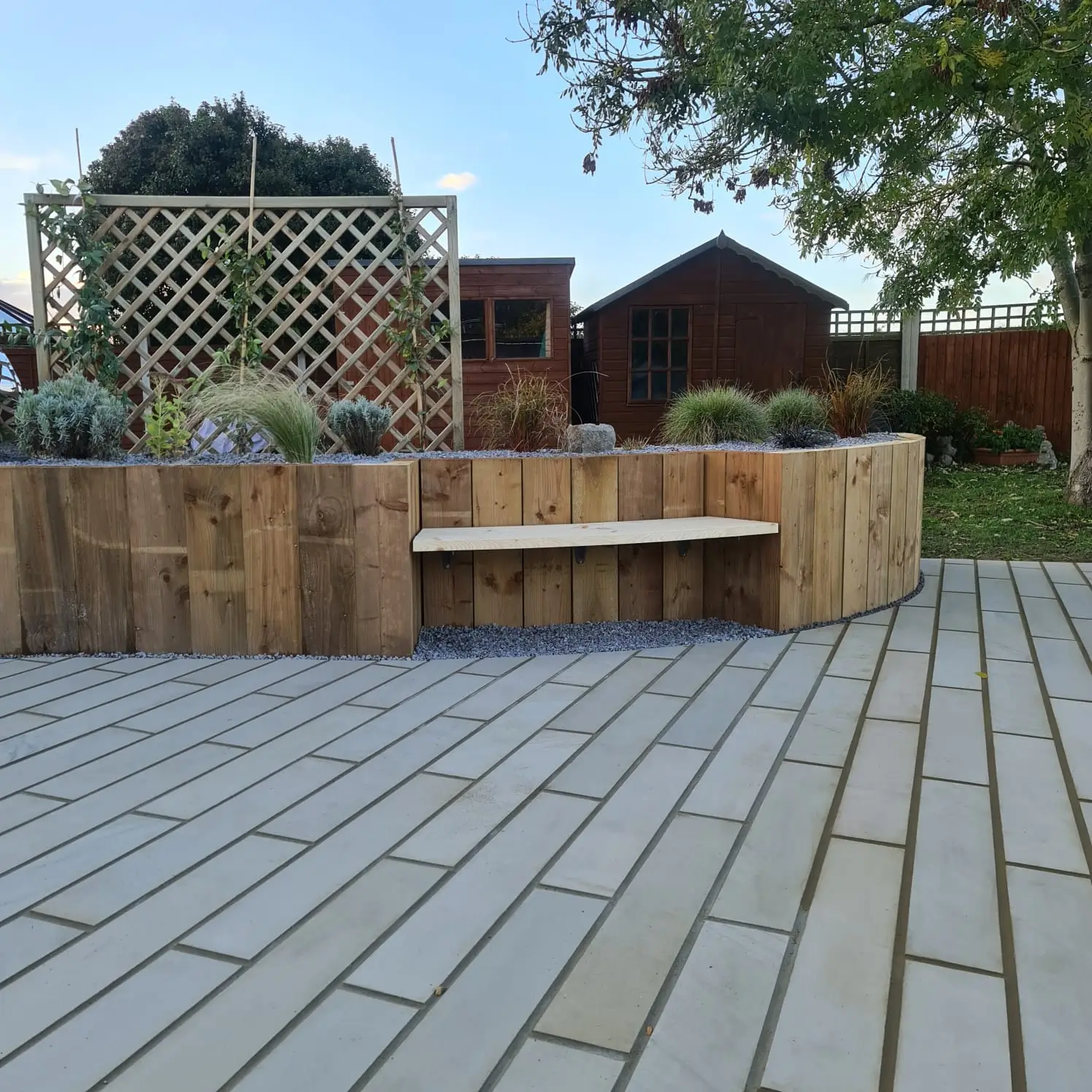 A patio with gray stone pavers, featuring a wooden seating area and greenery, set in a garden landscape.