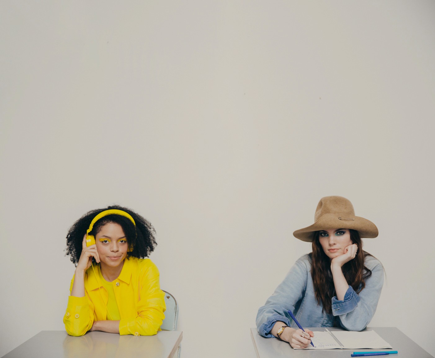Two women sit at desks in yellow and blue outfits, contrasting in style and expression