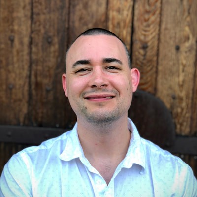 An image of Tim Preut facing the viewer with a smile and short hair, mid-thirties, with a wood backdrop and an iPhone portrait photo. 