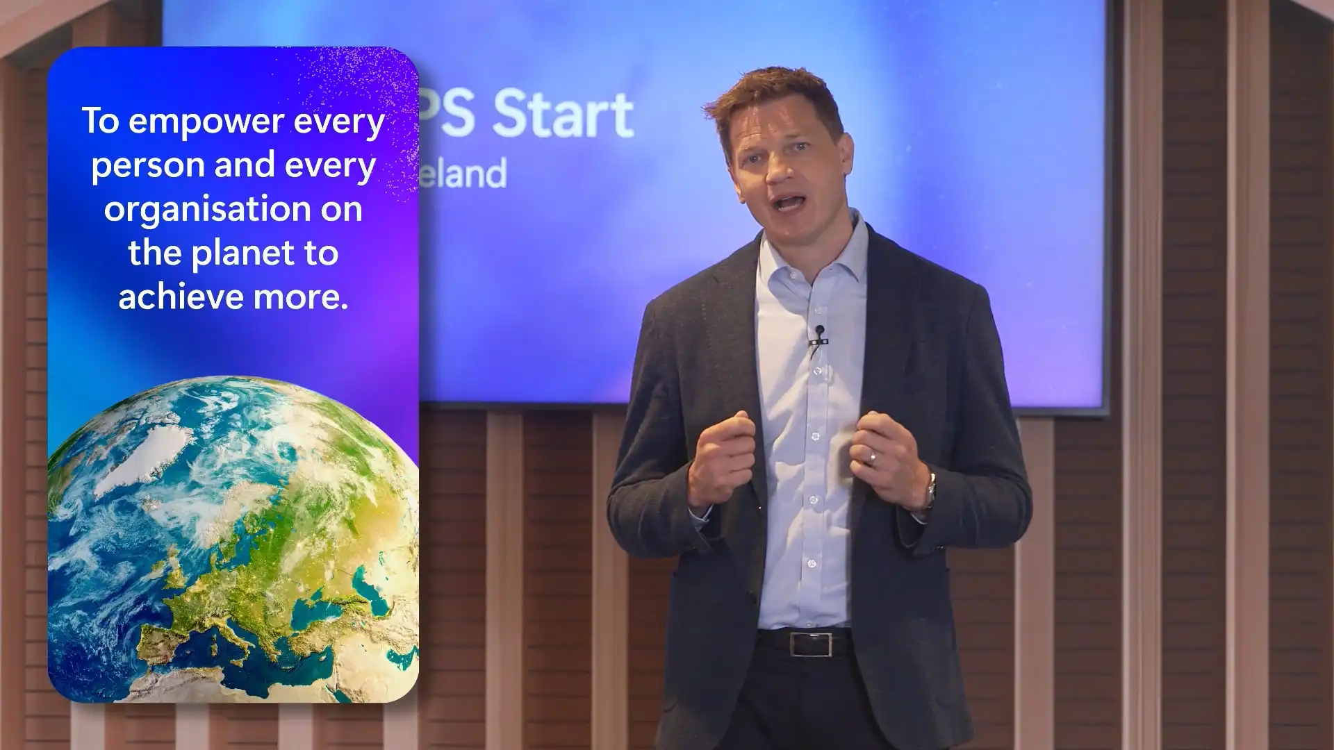 Presenter delivering a keynote-style message on stage alongside a large screen displaying a mission-led statement.