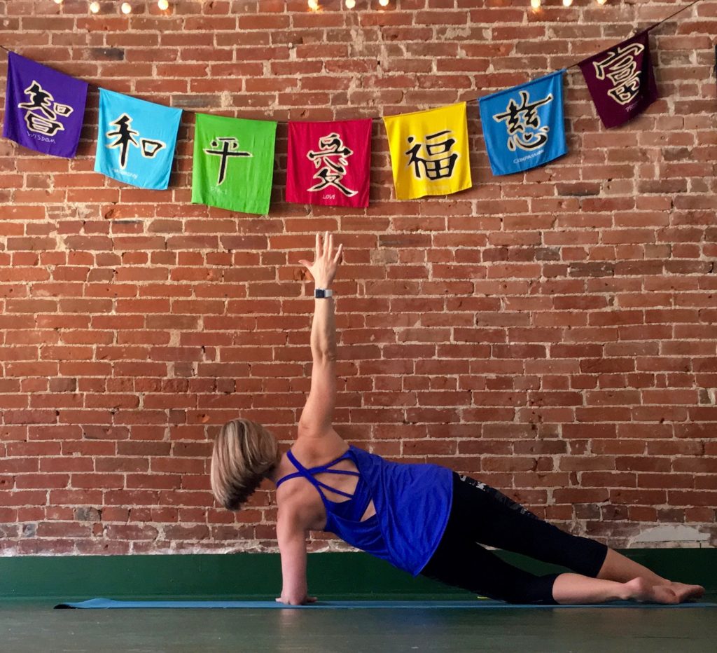 Heather Hausman in a blue tank top performs a side plank exercise on a turquoise yoga mat against a rustic brick wall.
