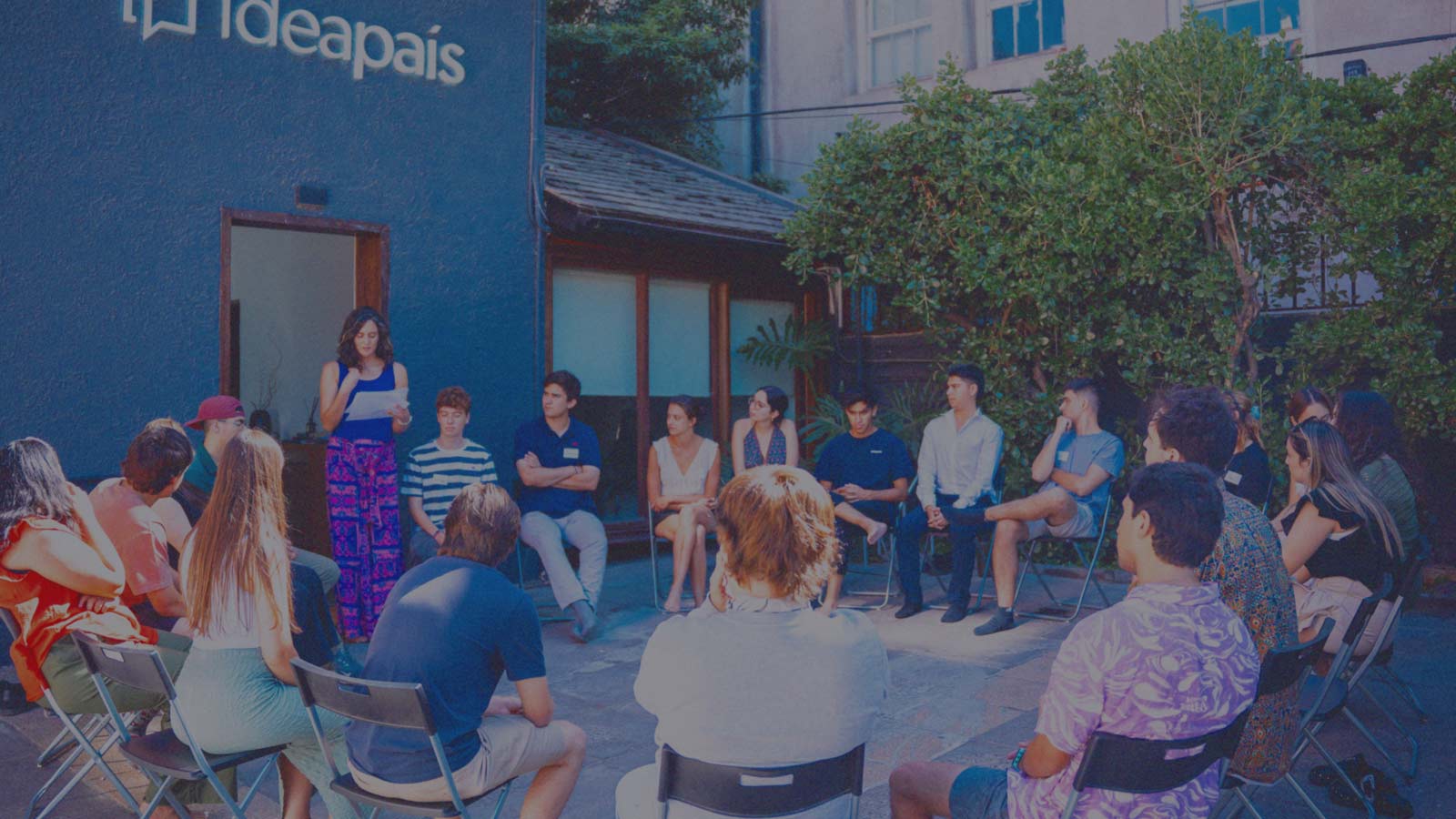 Portada de Colectivos de formación universitarios, programa de formación de IdeaPaís, mostrando a jóvenes sentados en un círculo en las afueras de la oficina de IdeaPaís mientras escuchan la charla que ofrece una investigadora. 