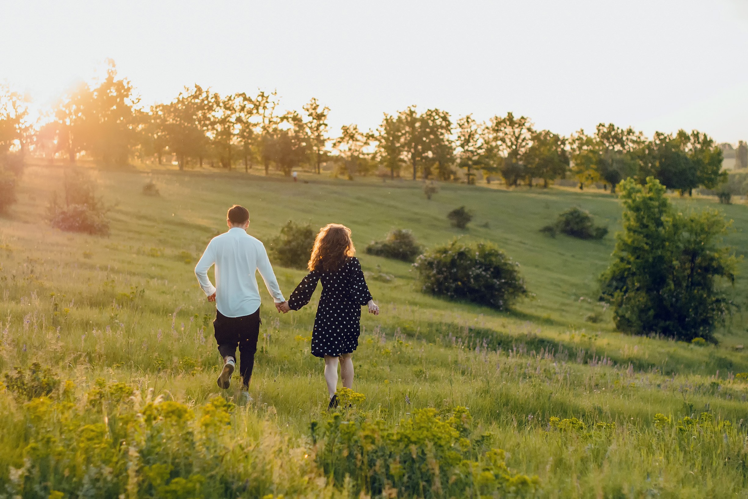 A couple walks hand in hand through a sunlit meadow.