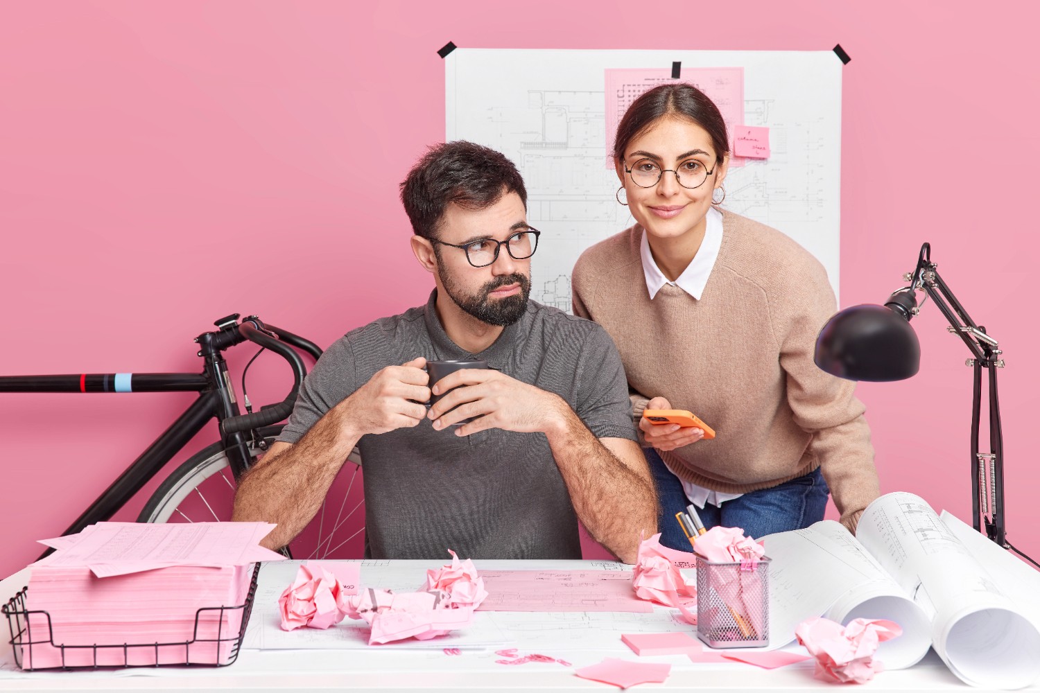 A man and woman collaborate at a desk, surrounded by pink paper, focused on their work.