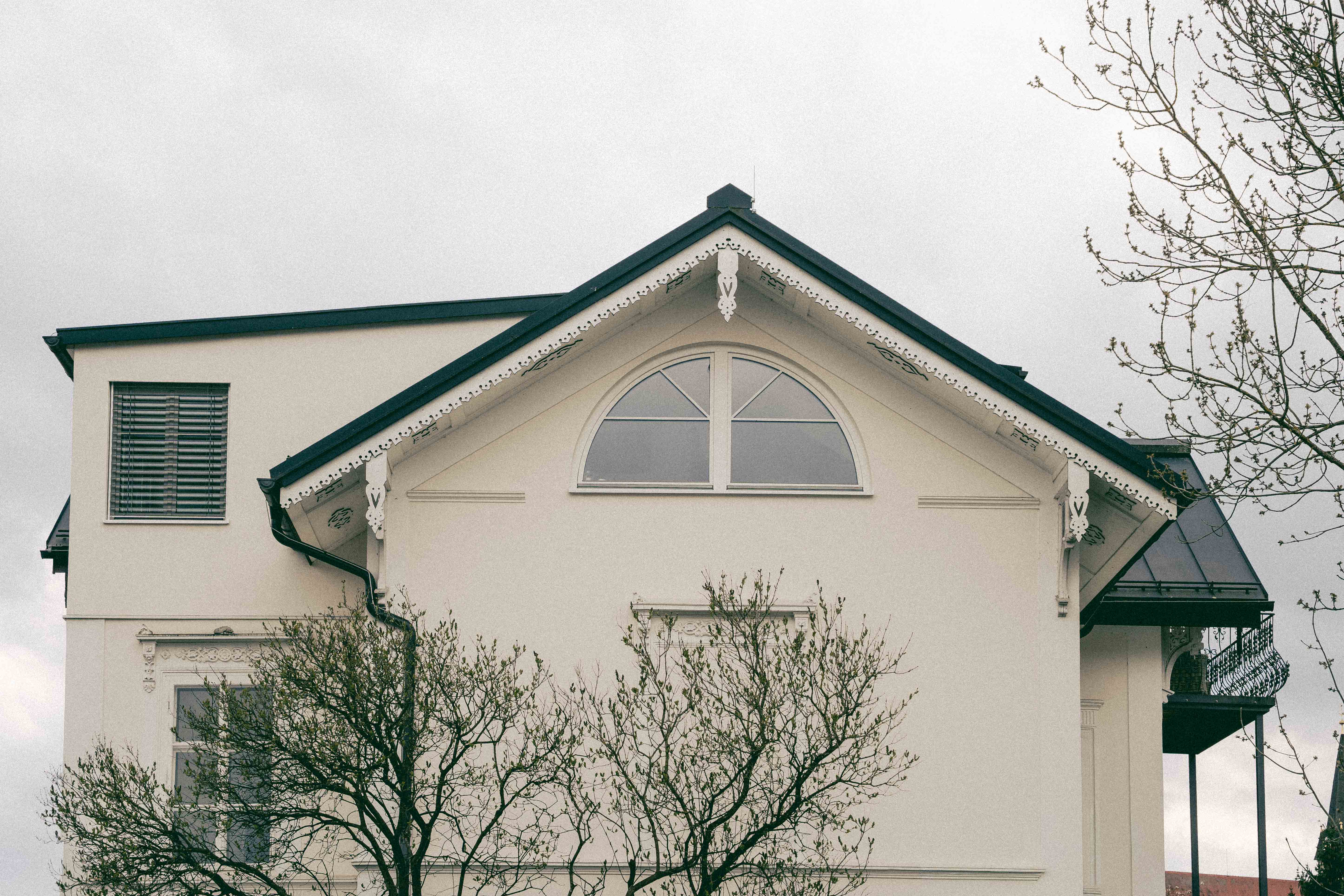 Ein weißes Haus mit einem halbkreisförmigen Fenster und einem schwarzen Dach. Frühling und grüne Bäume im Vordergrund.