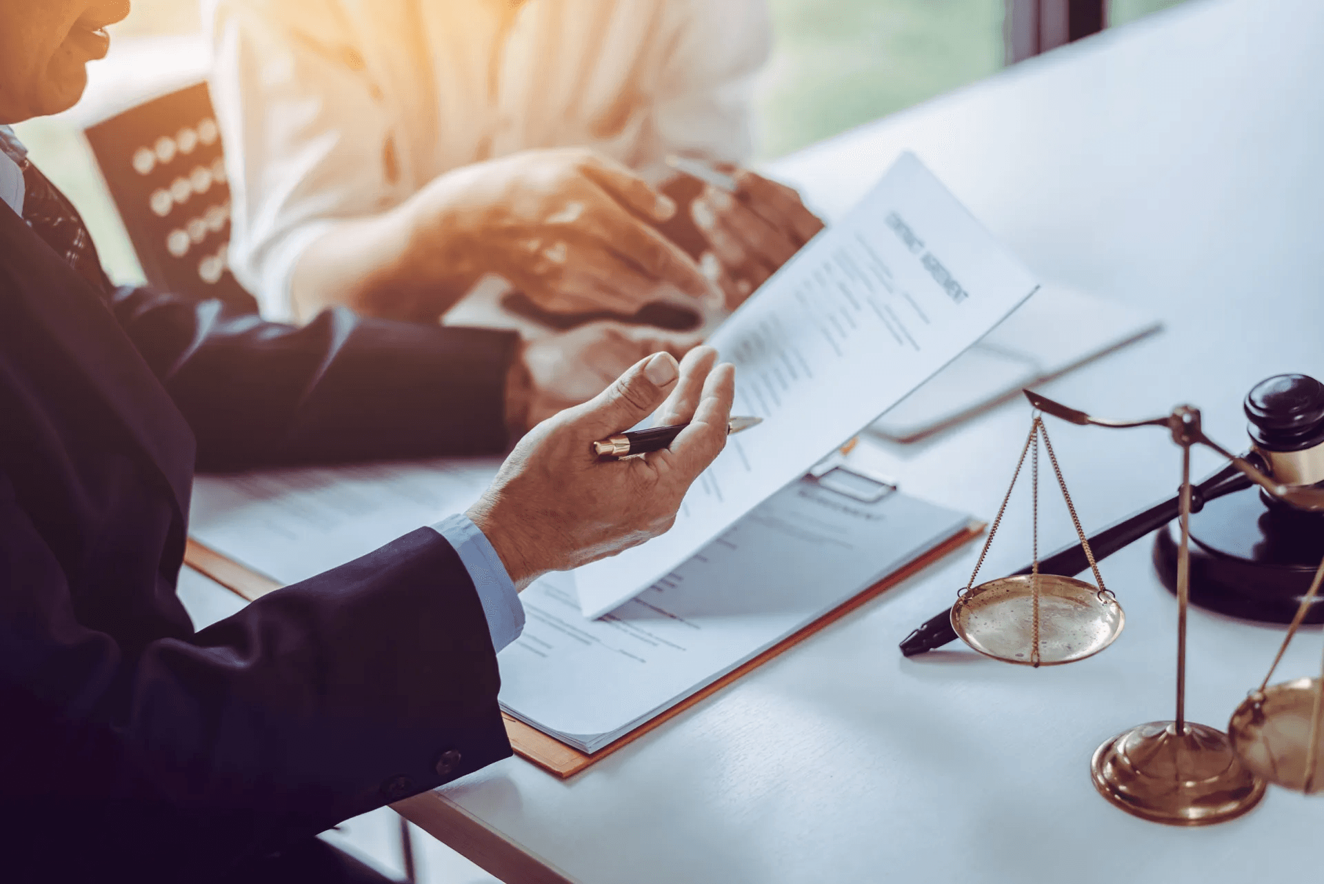 A business meeting scene with contracts on the table, one person holding a document while the other gestures, and a justice scale and gavel in the foreground.