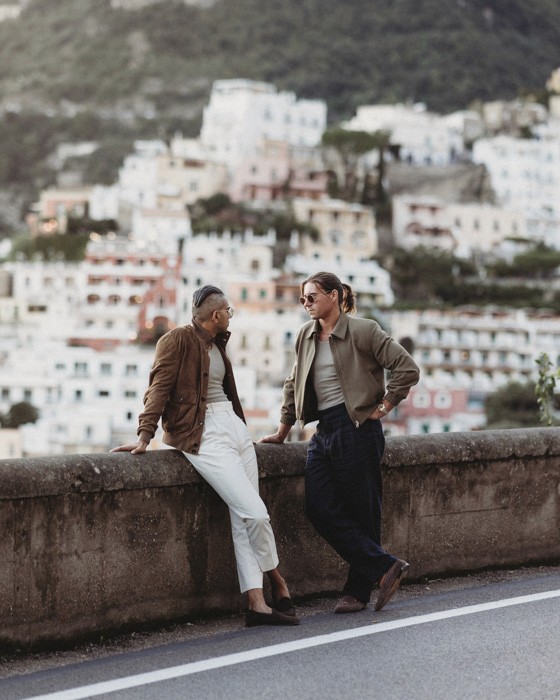 Two men in tailored jackets share a moment along the Italian coastline, showcasing the relaxed sophistication of Don Marrón.