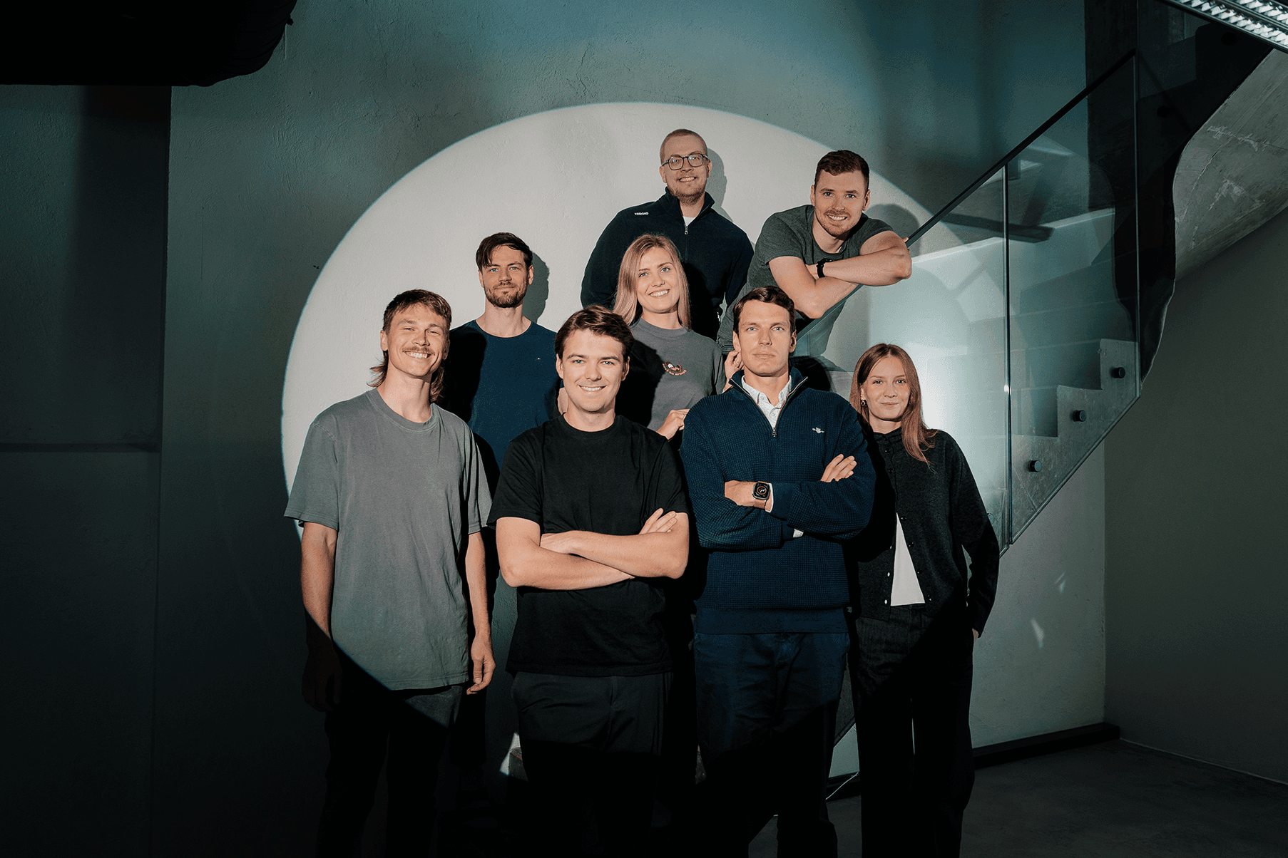 A team of eight sales outreach experts stands together inside a modern, industrial-style building, with glass railings and concrete stairs, under warm ambient lighting, conveying a sense of camaraderie and professionalism.