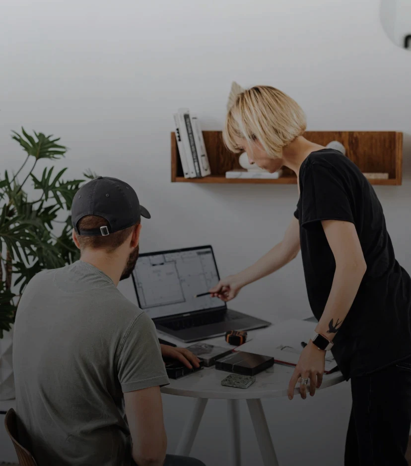 imagem de dois arquitetos trabalhando conjuntamente. Homem arquiteto está sentado olhando para o notebook. Mulher arquiteta está mostrando algo para o arquiteto no notebook.