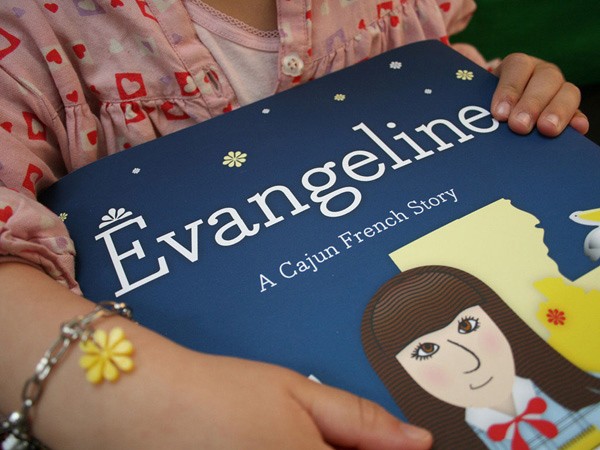 A child wearing a pink shirt with a pattern of hearts holds a dark blue book titled “Evangeline: A Cajun French Story.” The book cover features a cartoon illustration of a girl with brown hair and a red bow. The child’s left wrist has a yellow flower charm, and a silver bracelet with a small yellow flower charm is visible. The background is blurred and dark.