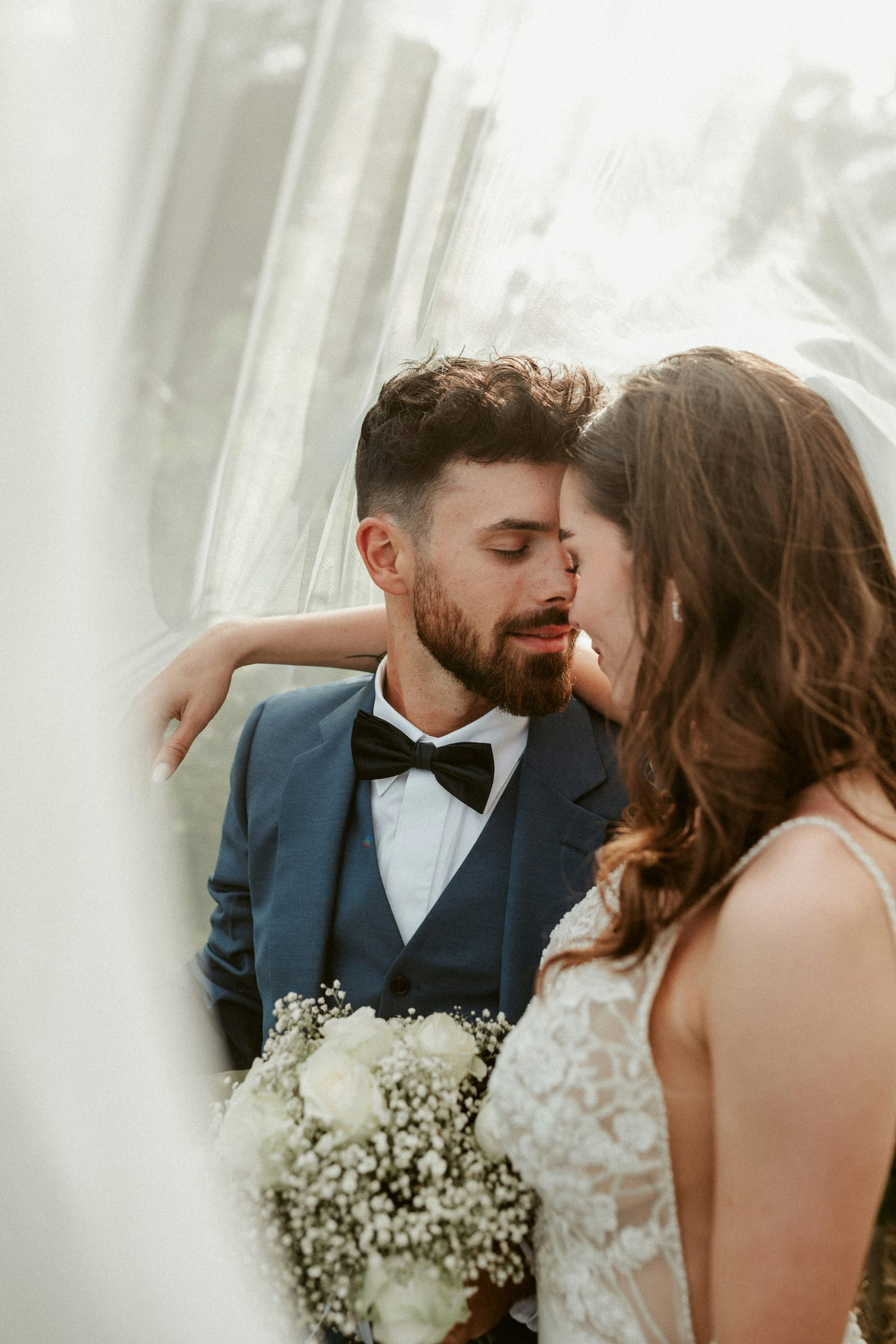 Intimate close-up of bride and groom beneath a sheer veil with a white rose bouquet and soft natural light