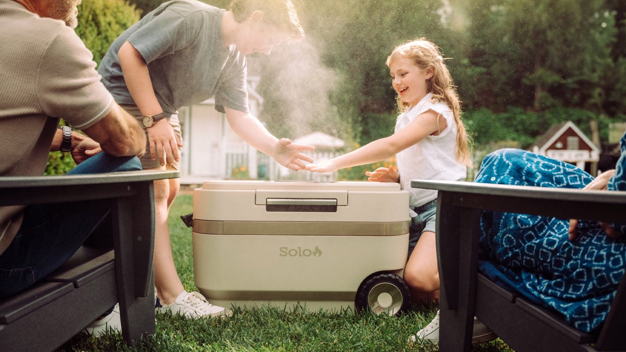 Two children reaching into mist rising from a cream wheeled cooler, set between two chairs on a backyard lawn