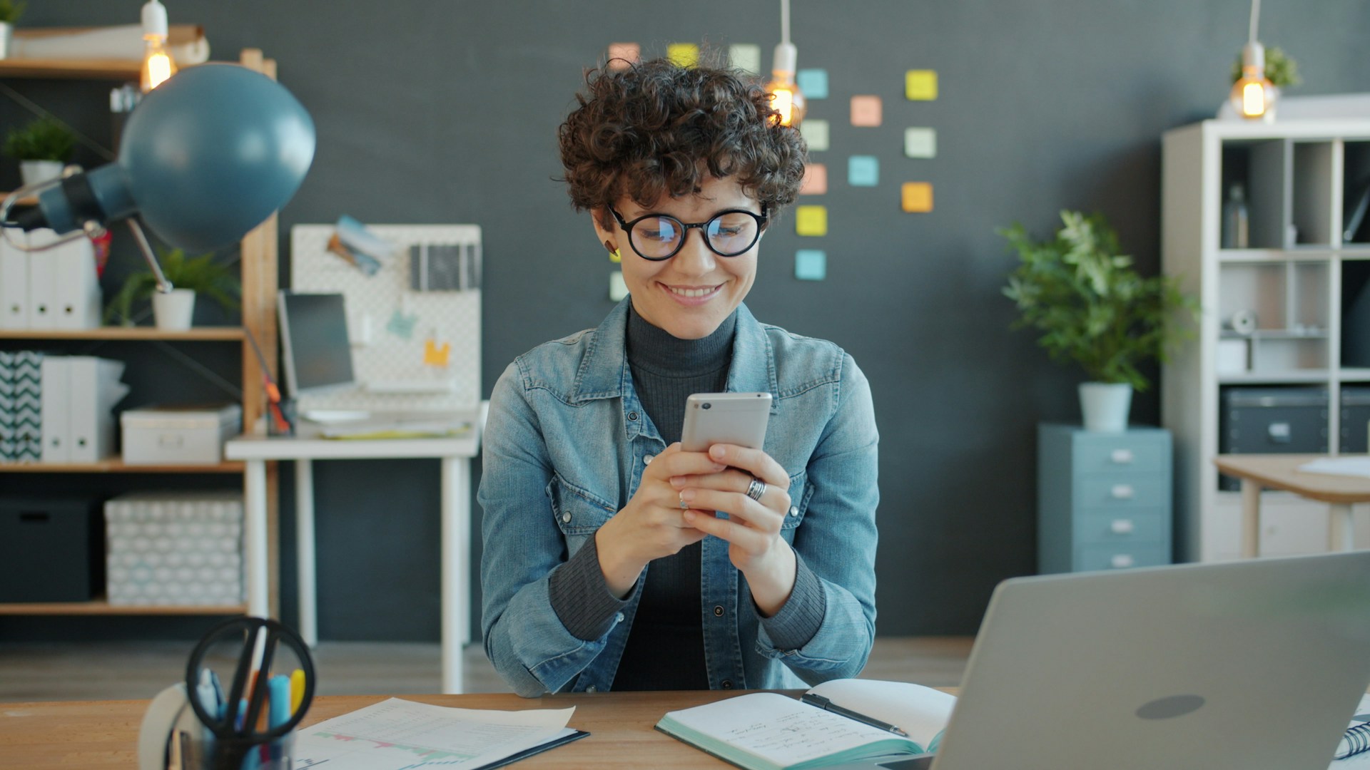 Smiling woman in glasses talking on a smartphone while looking at her laptop.