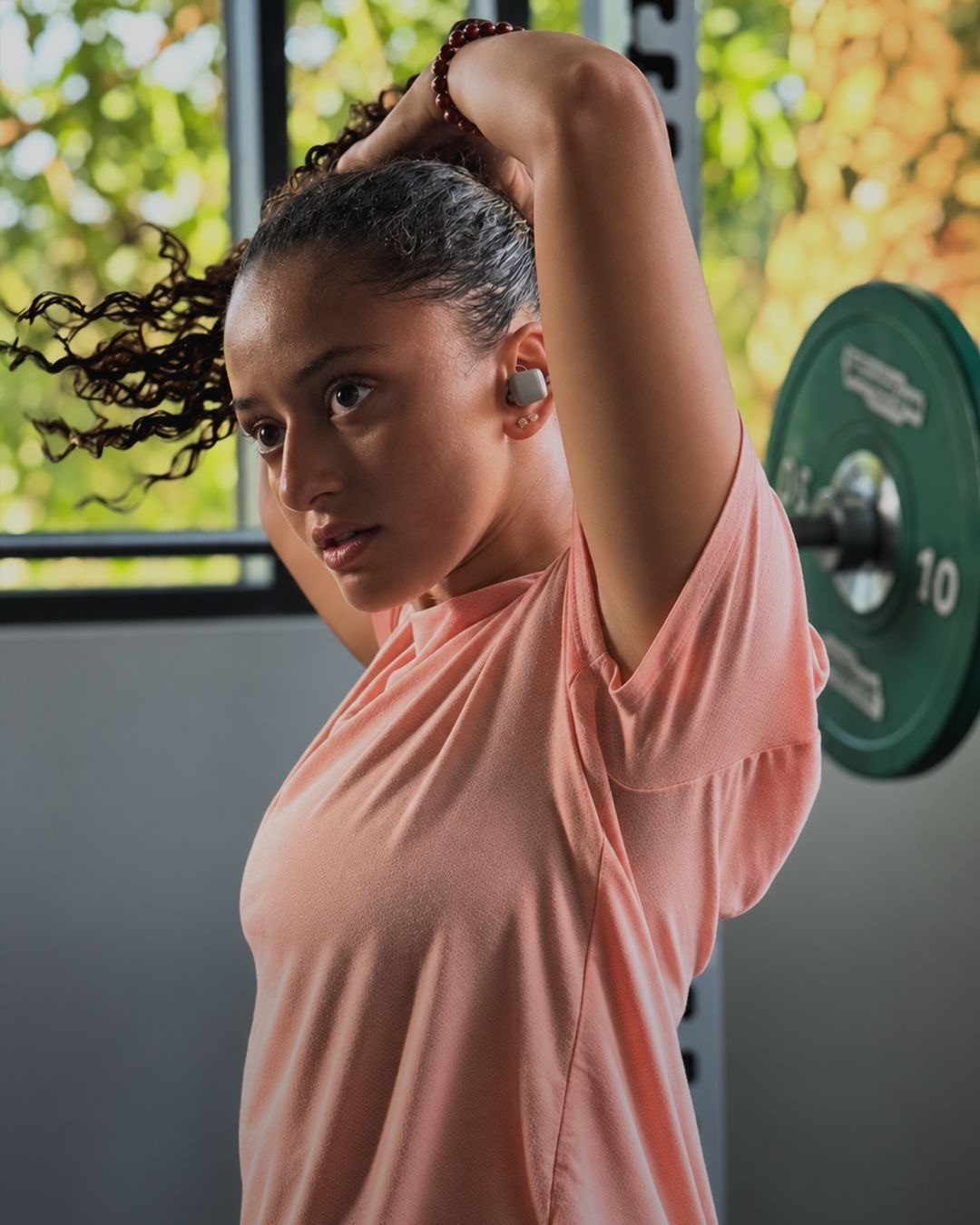 Lujain Khalil tying her hair in the gym in a peach training top, a loaded barbell behind her