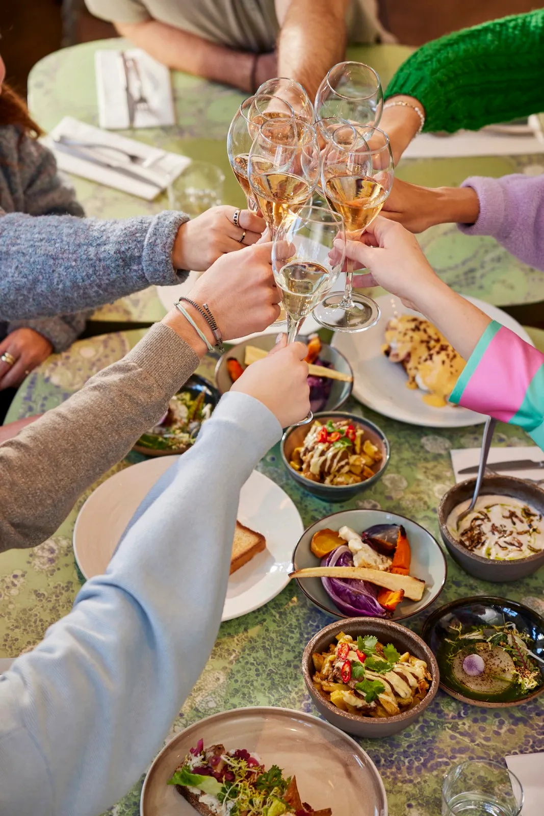 A top view of hands raising glasses for a toast, surrounded by plates of food on a table.