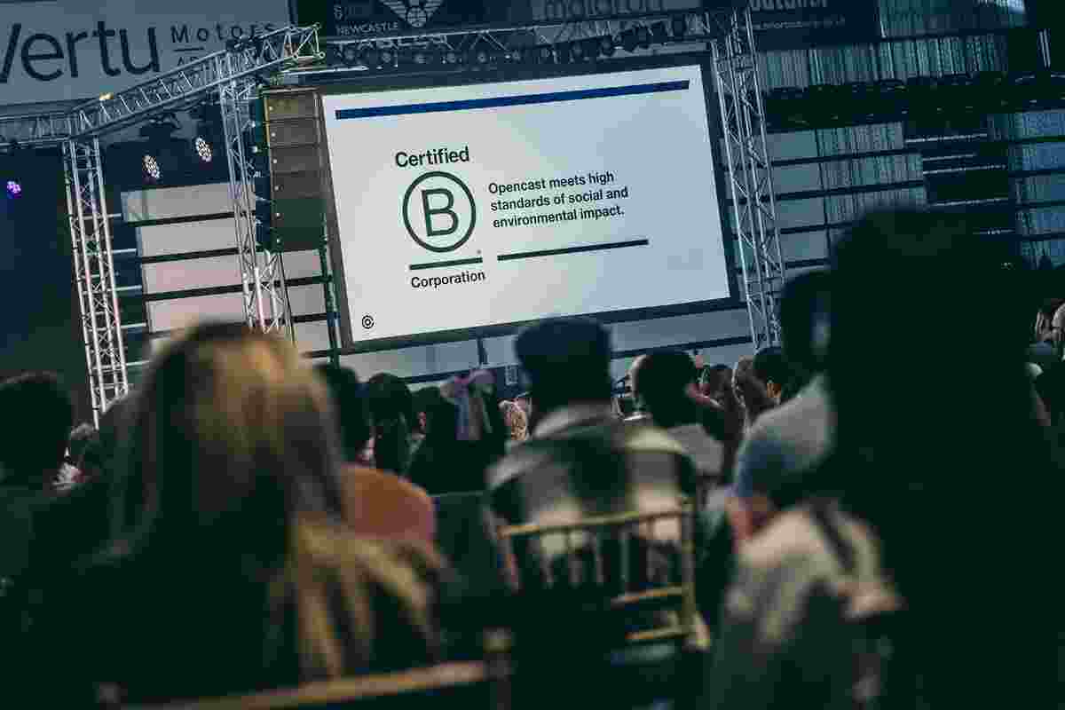 An audience seated in rows at a large indoor event, viewed from behind, facing a stage with a large presentation screen. The screen displays a “Certified B Corporation” logo alongside text stating that Opencast meets high standards of social and environmental impact. Stage lighting rigs and truss structures frame the screen, and the surrounding space resembles a conference or award‑style venue. The scene conveys a formal announcement or presentation during a professional gathering.