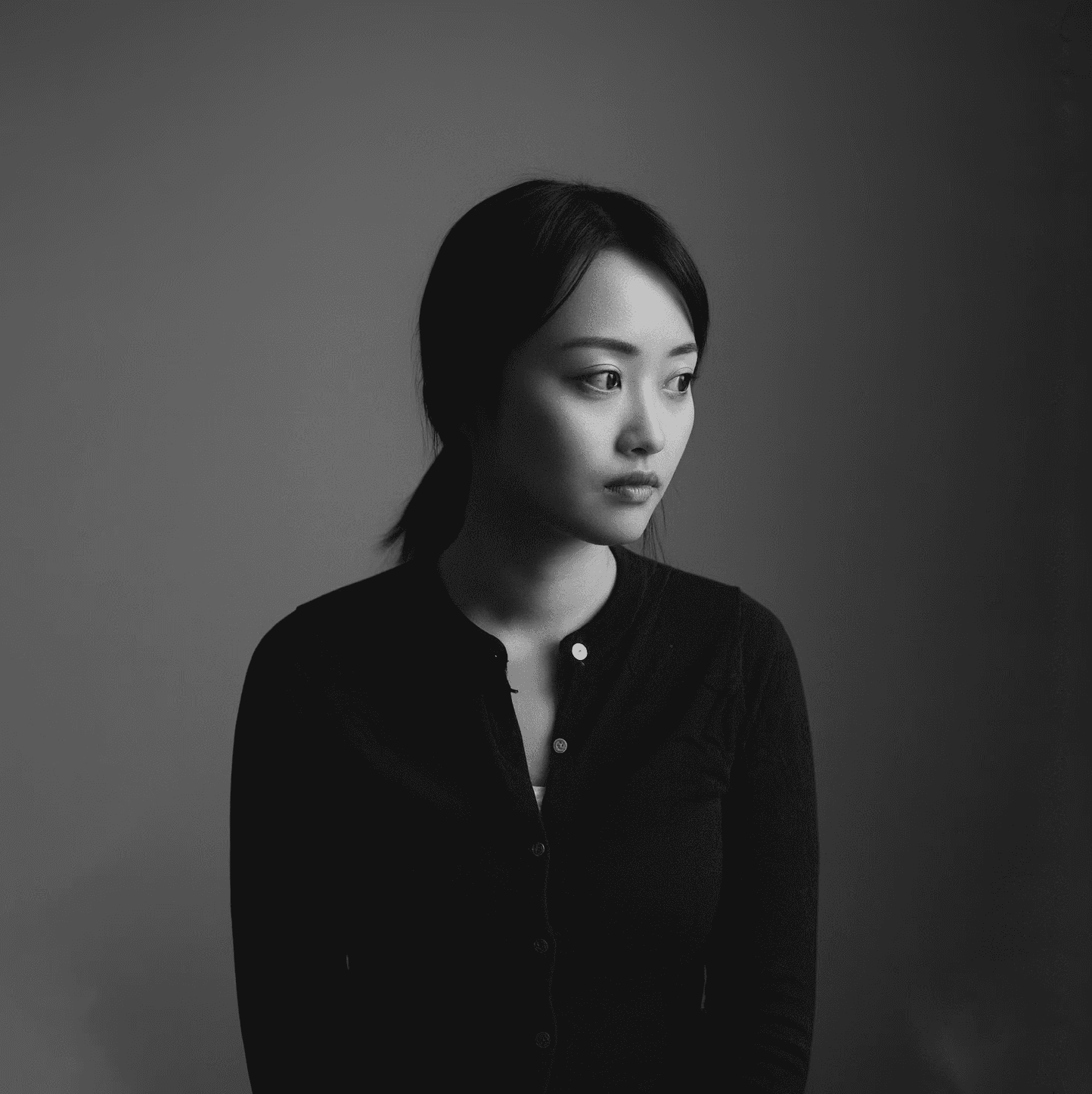 A thoughtful young person with dark hair gazes to the side, captured in soft black and white lighting.