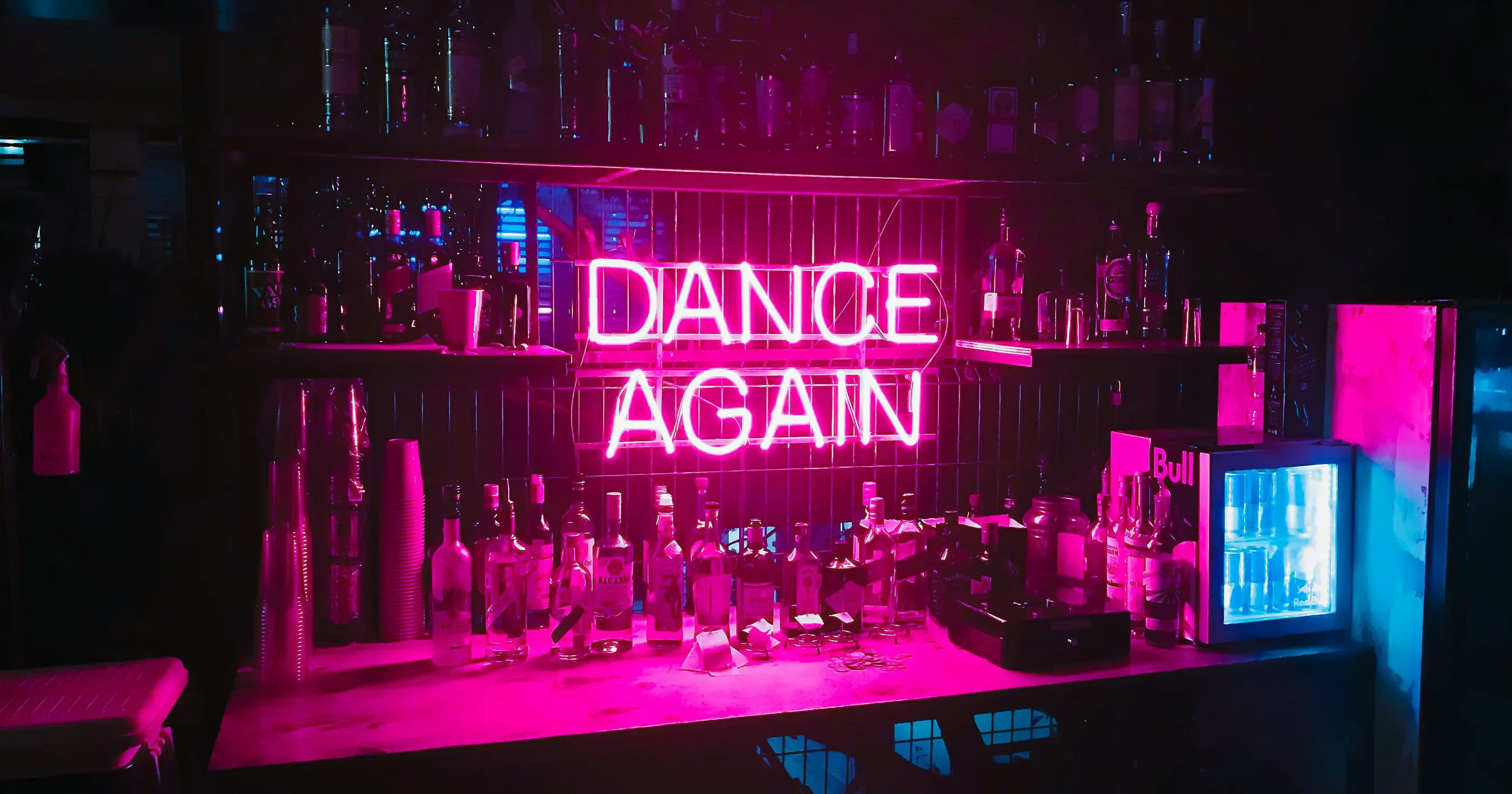 Bar with a pink neon ‘Dance Again’ sign, liquor bottles, and moody club lighting.