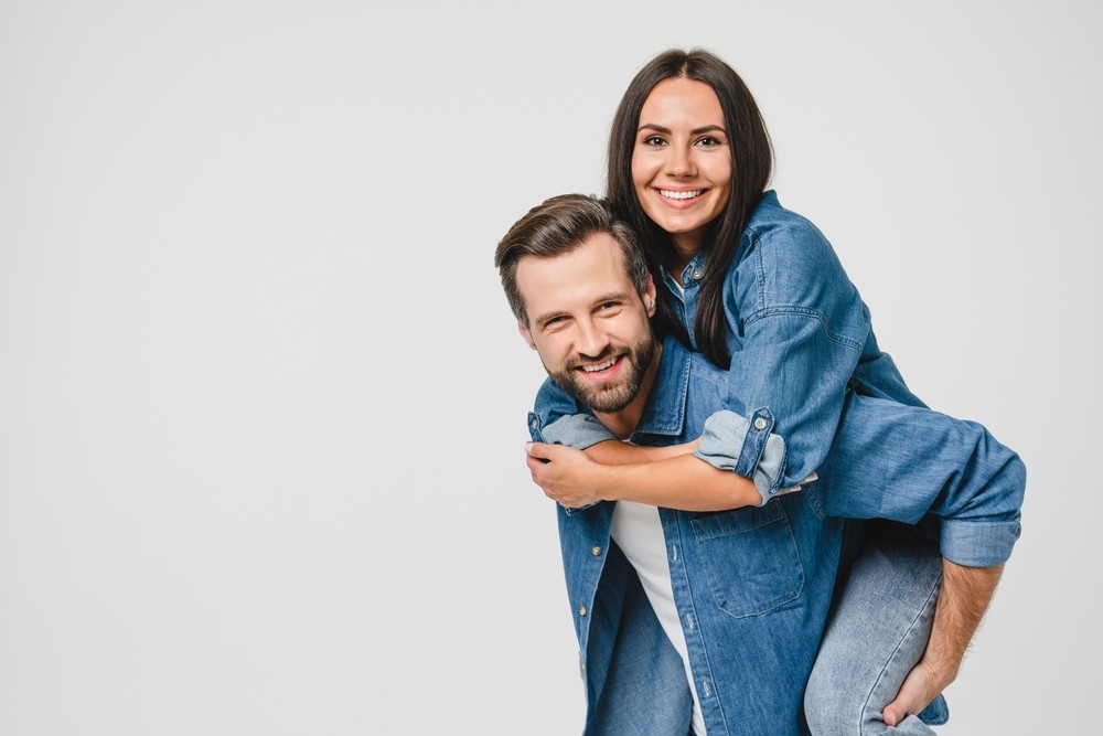 Two people are doing a piggyback couple pose in matching denim outfits against a white background.