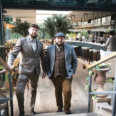 Two men, Adam Yeomans and Ben Fox of Hartingtons Heritage Tours, in Derbyshire. They wear tweed suits in brown and grey and one wears flat cap walking up stairs