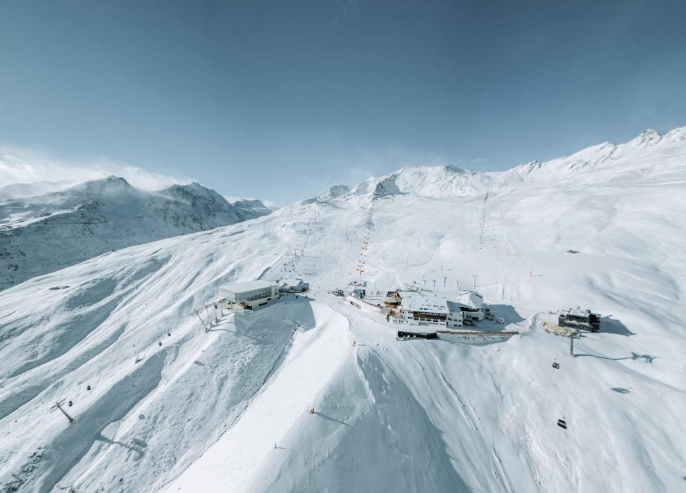 Luftaufnahme vom schneebedeckten Bergresorts Sölden mit Skiliften, umgeben von weitläufigen schneebedeckten Gipfeln unter einem klaren blauen Himmel.