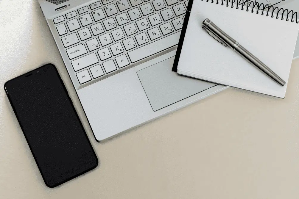 Laptop keyboard with a notebook, pen, and smartphone on a desk.