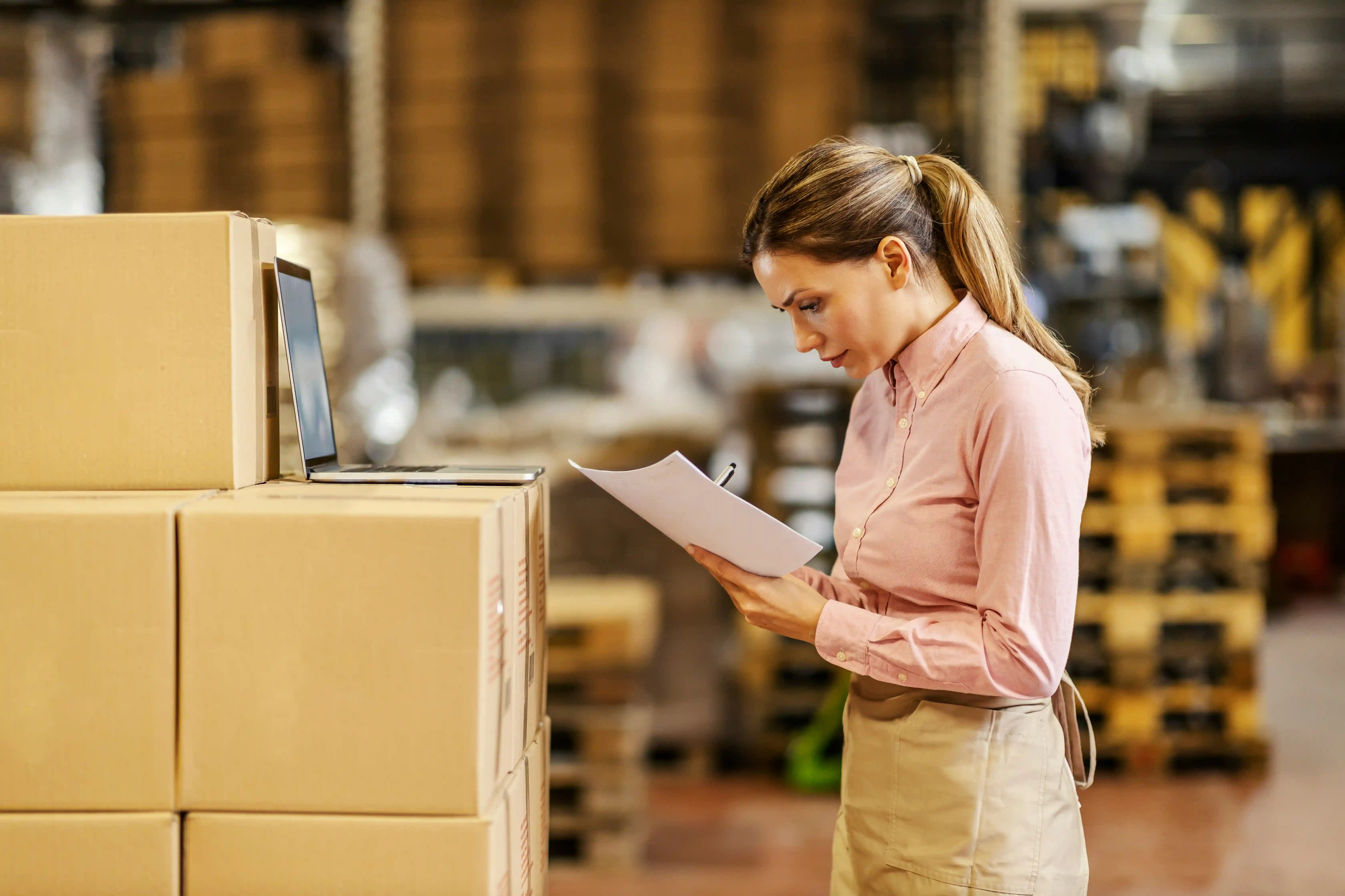 woman stands in warehouse withj laptop and paper