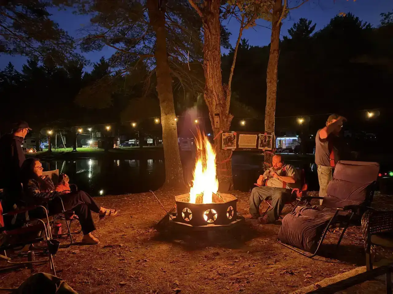 People sitting around a star-patterned fire pit at night with a view of the pond at Pine Hollow Campground in Southern Vermont.