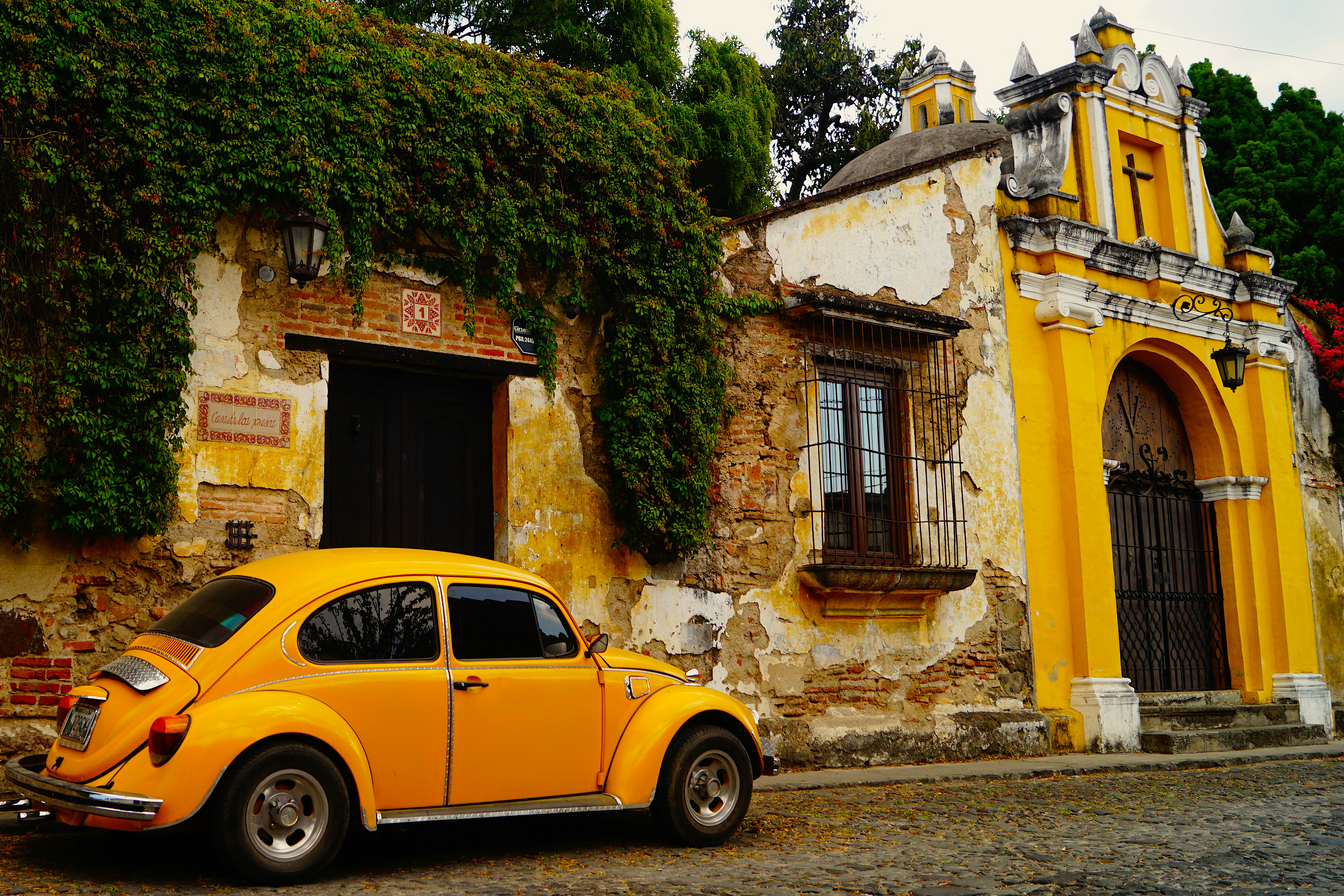 A vibrant yellow vintage Volkswagen Beetle is parked in front of an old, ivy-covered building with crumbling walls and a distinctive yellow archway, capturing a nostalgic scene on a cobblestone street.