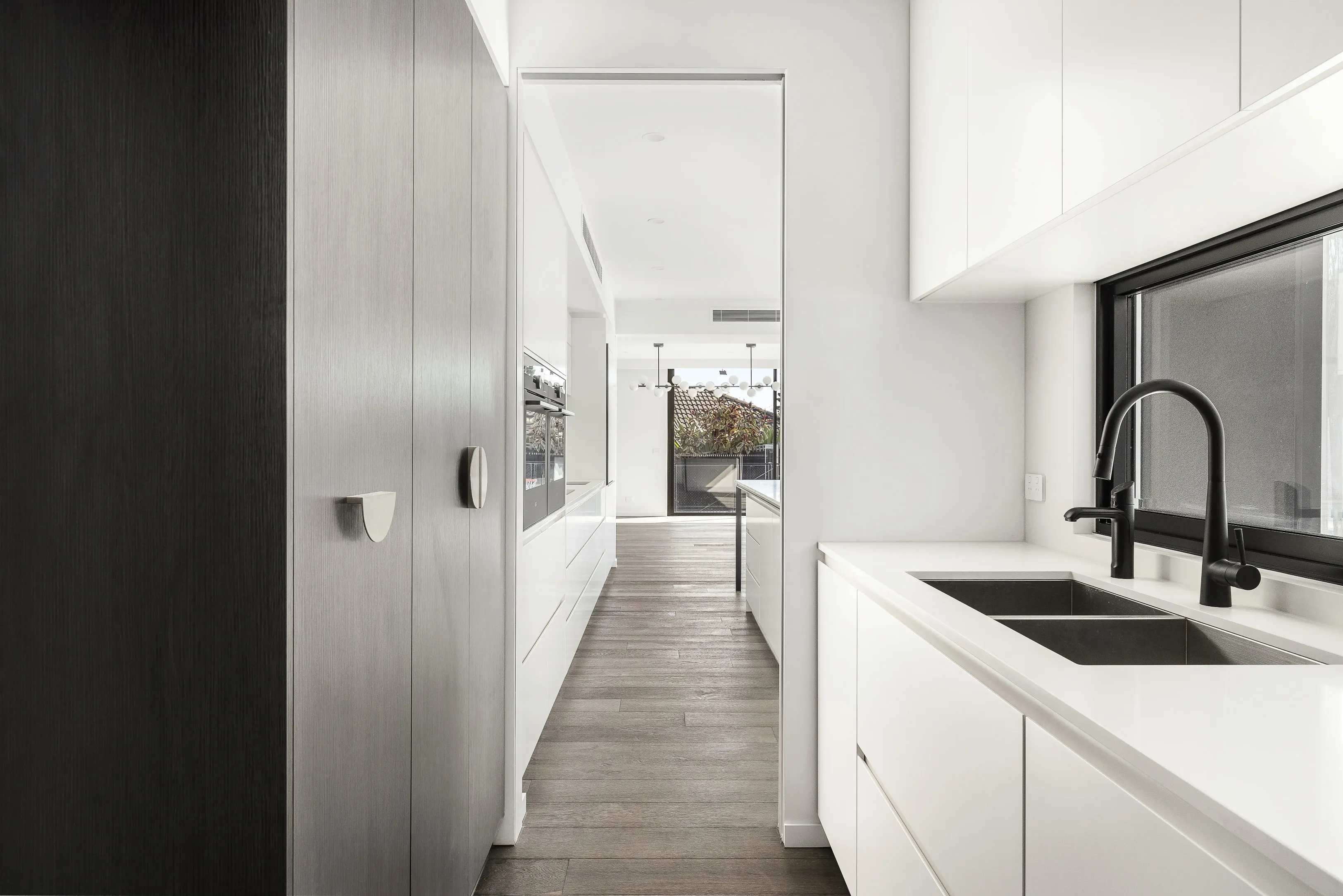 Butler's pantry with black and pale-timber tall cabinetry, white benchtop, black tap and sink and a window, looking through to the main living area