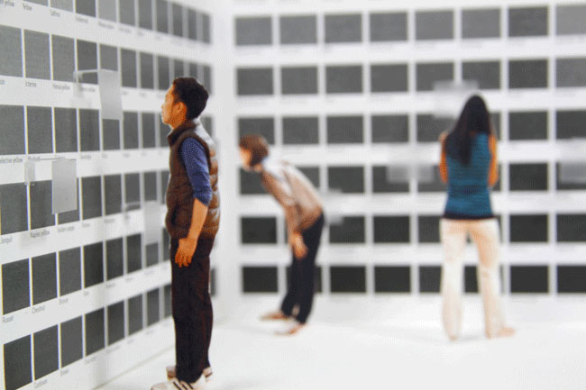Three people are in a room with a white wall displaying many small square images. The person on the left wears a brown vest and dark pants. The person in the center wears a light brown long-sleeved shirt and dark pants. The person on the right wears a teal shirt and light-colored pants. The floor is white, and the wall is white with evenly spaced square images.