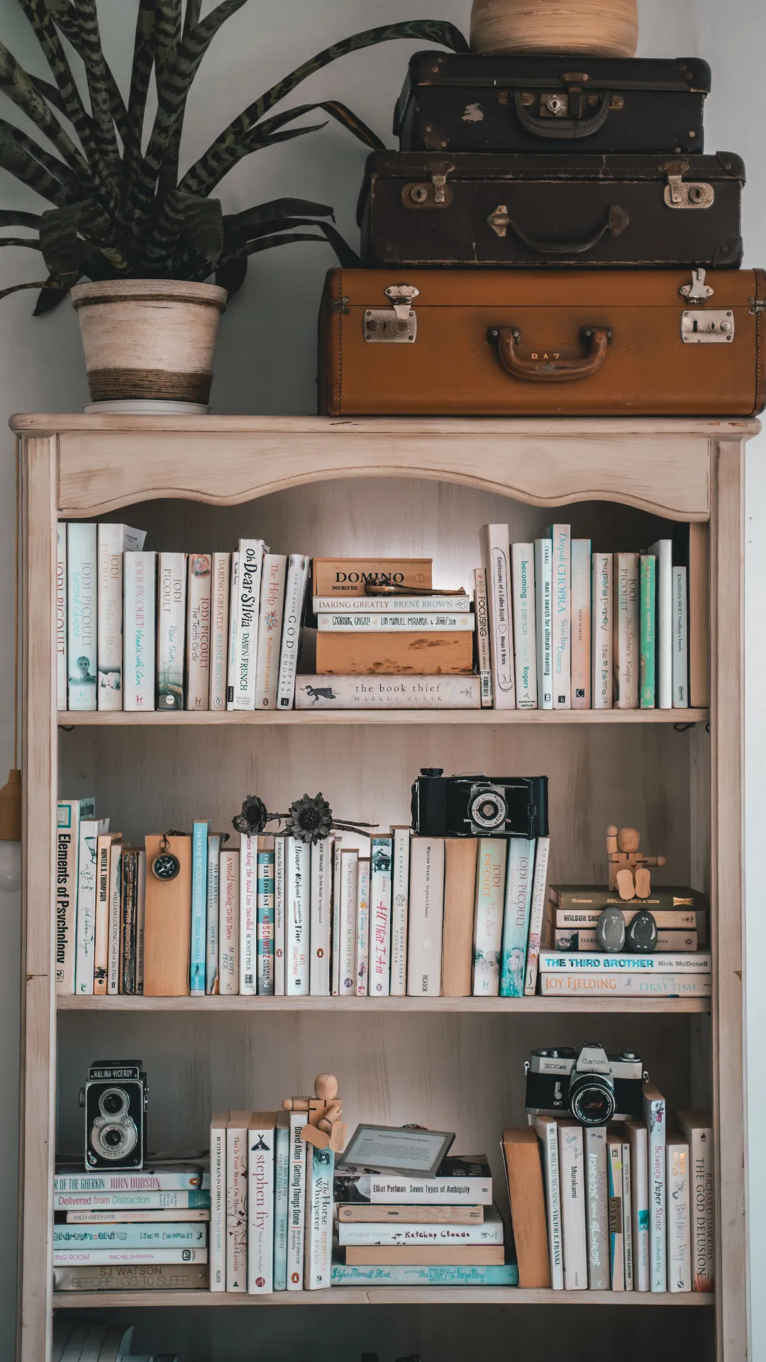 A wooden bookcase filled with various books and stacked suitcases at the bottom, creating a cozy reading corner.