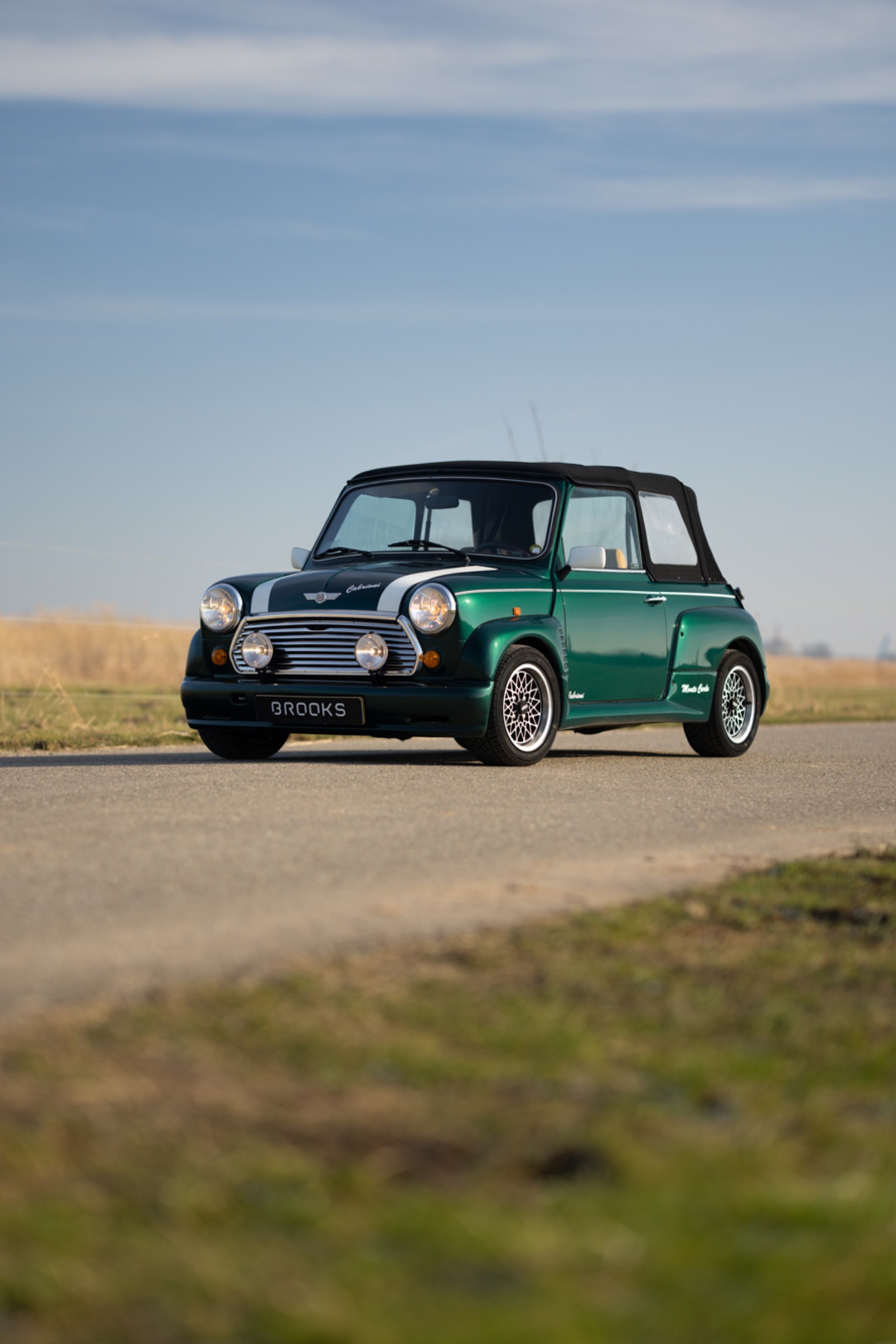 A classic green car parked on a road with open fields and a blue sky in the background.