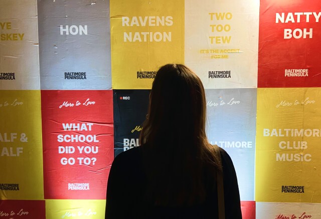 A person with long hair stands facing a wall covered in colorful tiles featuring various phrases and words related to Baltimore.