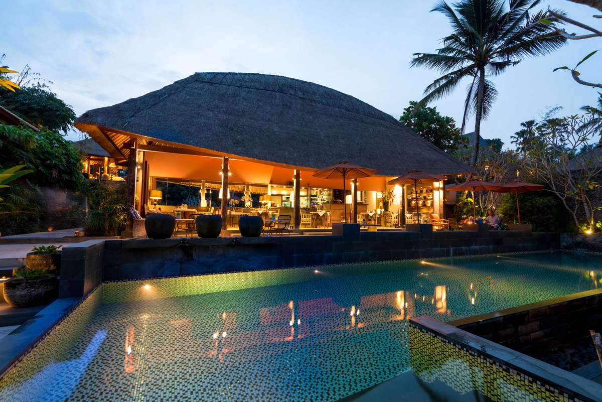 A thatched-roof tropical pavilion with a lit interior sits behind an illuminated turquoise swimming pool at dusk.