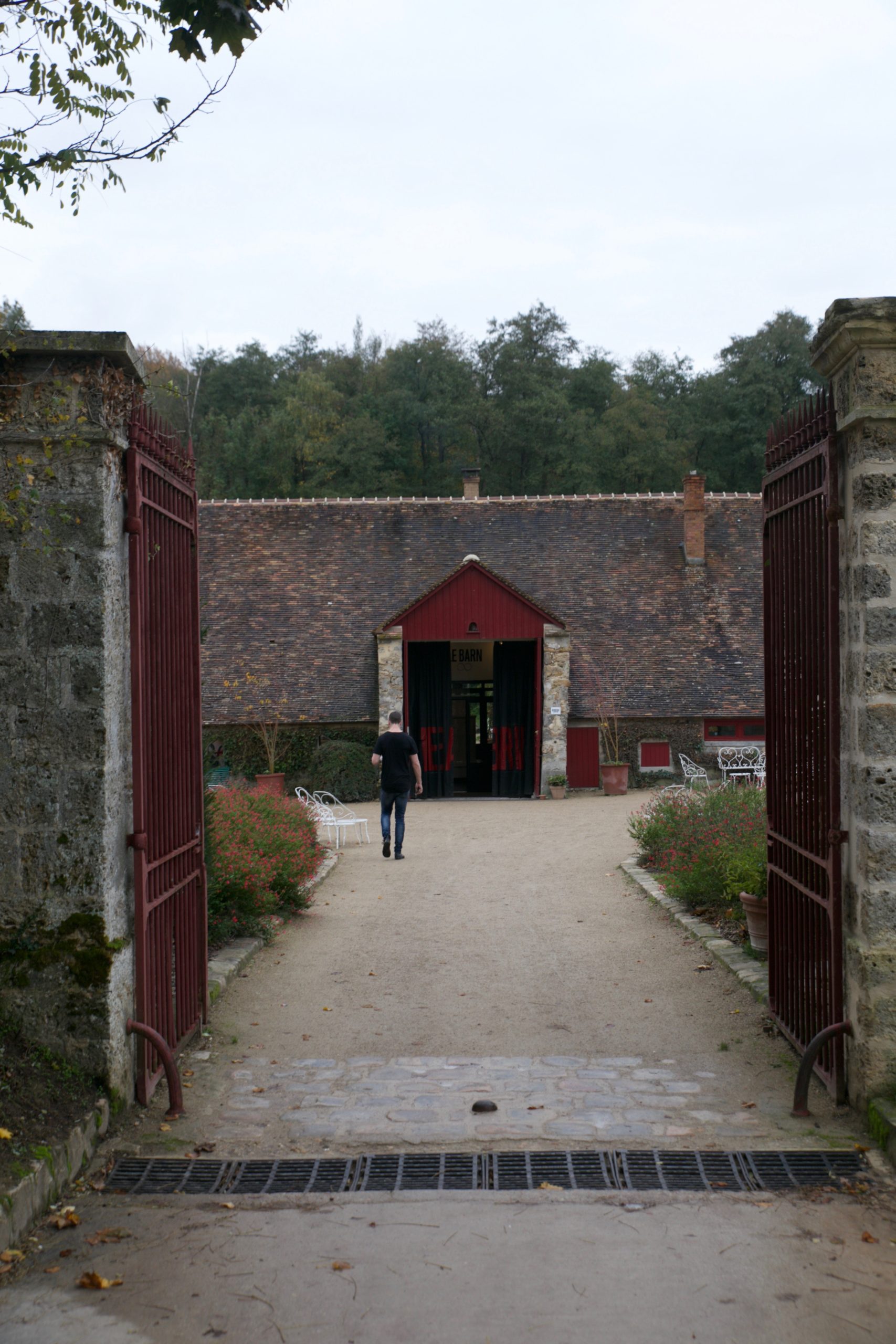 Open red iron gates leading up a gravel path to a stone barn with a canvas "LE BARN" sign